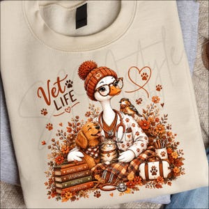 Puede incluir: Una sudadera beige con un gráfico de un ganso con gorro, gafas y estetoscopio. El ganso está rodeado por un perro, un gato, un pájaro pequeño, una pila de libros con las etiquetas Animal Care, Veterinary, Health, Compassion y flores.