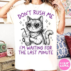 May include: Cream-colored t-shirt with a graphic of a cat wearing heart-shaped sunglasses and a bandana, holding a bubble tea. The text reads "DON'T RUSH ME I'M WAITING FOR THE LAST MINUTE" in purple.