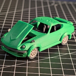 May include: A bright green model car with its hood open, revealing the engine compartment. The car has a classic sports car design with a spoiler and detailed wheels. The model is set against a grid background.
