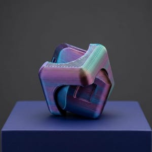 May include: A 3D printed geometric puzzle cube featuring a multi-coloured iridescent finish that shifts between shades of blue, purple, and teal. The object displays visible horizontal layer lines from the printing process and has a complex, interlocking structure with rounded corners and recessed square panels on its faces.