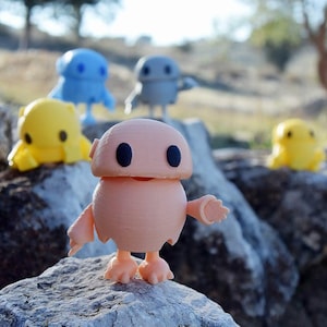 Puede incluir: Una colección de figuras impresas en 3D en varios colores, incluyendo melocotón, azul, amarillo y gris. Las figuras tienen un diseño redondeado con ojos grandes y ovalados y brazos y pies pequeños. Están posadas sobre una superficie rocosa con un fondo exterior borroso.