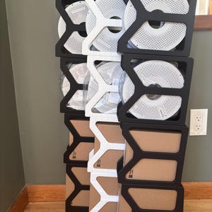 May include: A vertical stack of modular storage organizers in black and white, holding circular 3D printer filament spools and square cardboard boxes. The organizers feature a Y-shaped structural frame design, creating a grid for storage.