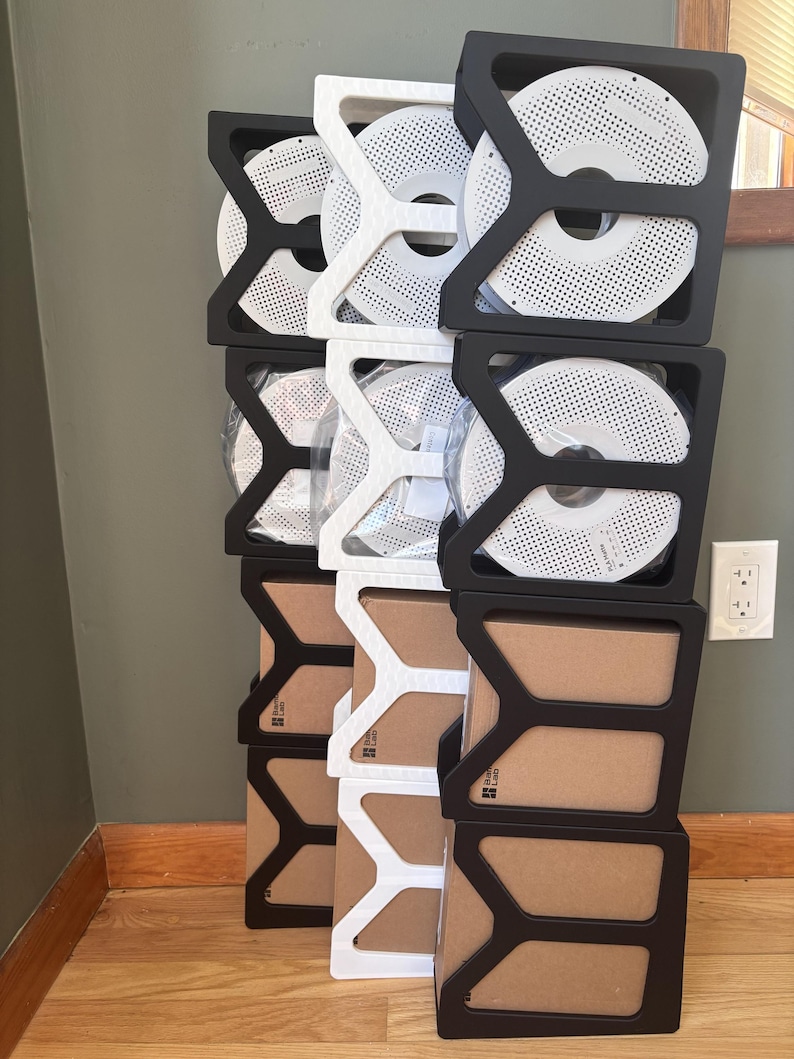 May include: A vertical stack of modular storage organizers in black and white, holding circular 3D printer filament spools and square cardboard boxes. The organizers feature a Y-shaped structural frame design, creating a grid for storage.