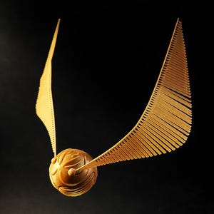 May include: A golden Snitch, a fictional object from the Harry Potter series, is suspended against a black background. The Snitch features a detailed, textured sphere with two sets of delicate, wing-like structures extending outwards.
