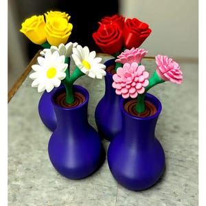 Puede incluir: Cuatro jarrones azul oscuro, cada uno con un tipo de flor diferente: rosas amarillas, rosas rojas, margaritas blancas y claveles rosas. Los jarrones están dispuestos sobre una superficie gris claro. Arreglo floral decorativo.