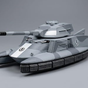 May include: A gray and white toy tank with a long barrel and a distinctive design. The tank features a camouflage pattern and a white stripe with a circular emblem. The toy tank is a model of a military vehicle.