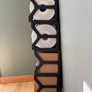 May include: A vertical stack of four black modular storage frames. The top two frames hold white circular filament spools with perforated patterns. The bottom two frames contain brown cardboard boxes, with one box displaying the Bambu Lab logo.