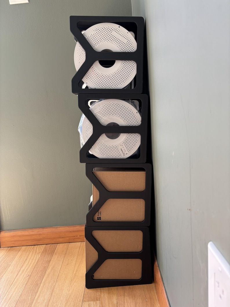 May include: A vertical stack of four black modular storage frames. The top two frames hold white circular filament spools with perforated patterns. The bottom two frames contain brown cardboard boxes, with one box displaying the Bambu Lab logo.