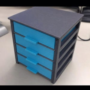 May include: A small, four-drawer storage unit. The unit is primarily gray with four blue drawers. Each drawer has a small, rectangular blue handle. The top and sides of the unit are gray. The unit is designed to sit on a desk or table.
