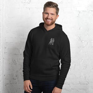 Może przedstawiać: Czarna bluza z kapturem z przednią kieszenią i białym obrysem aparatu i dwóch postaci. Bluza wykonana jest z miękkiego materiału.