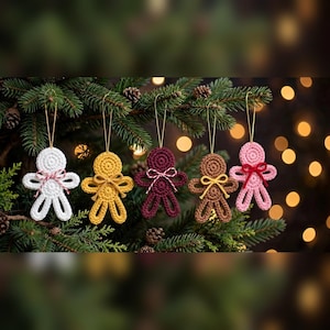 May include: Five crocheted gingerbread-style ornaments hanging from a pine tree. The figures are white, yellow, dark red, brown, and light pink. Each ornament features a small bow at the neck and a gold string loop for hanging on a branch.