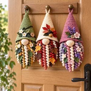 Peut inclure: Trois décorations de gnomes en crochet suspendues avec des chapeaux pointus. Le gnome de gauche est vert avec des fleurs blanches, celui du centre est crème avec des feuilles d'automne et des baies, et celui de droite est mauve avec des fleurs violettes.