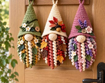 May include: Three hanging crochet gnome decorations with pointed hats. The left gnome is green with white flowers, the center is cream with autumn leaves and berries, and the right is mauve with purple flowers. Each has a round nose and curly beard.