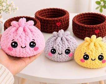 May include: Three crocheted plush dumplings in pink, light gray, and yellow, each featuring embroidered black eyes, a small smile, and pink cheeks. Behind them are three dark brown crocheted baskets, each decorated with a small red heart.