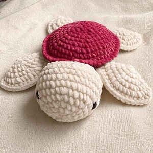 Peut inclure: Une tortue en peluche crochetée à la main avec une tête et quatre nageoires texturées de couleur crème en fil doux. La tortue possède une carapace arrondie proéminente d'un magenta profond et deux petits yeux ronds et sombres sur la tête.