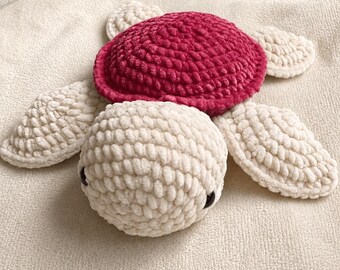 May include: A handmade crochet turtle plush toy featuring a textured, cream-colored head and four flippers made from soft yarn. The turtle has a prominent, rounded shell in a deep magenta color and two small, dark, round eyes on its head.