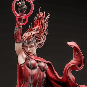May include: A detailed statue of a character in a red costume with flowing red hair. The figure is holding a glowing red ring above their head. The statue has a determined expression and is set against a dark background.
