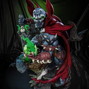 Puede incluir: Estatua detallada de Spawn, un personaje de cómic, con capa roja y armadura con púas. La figura sostiene un objeto verde brillante y una espada. La estatua está sobre un fondo oscuro y texturizado.