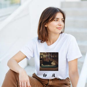 Può includere: T-shirt bianca con una grafica di un gatto nero su un letto in una stanza. La grafica include testo giapponese e un'illustrazione. La maglietta è indossata da una persona.