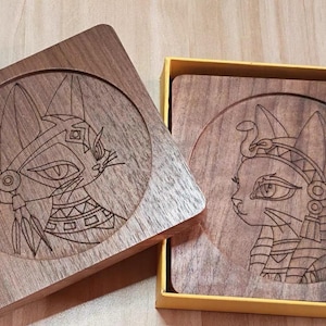 Puede incluir: Posavasos cuadrados de madera con diseños de gatos grabados. Uno presenta un gato con tocado, el otro un gato con corona de serpiente. Los posavasos están en una caja amarilla.