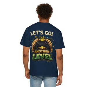 May include: Navy blue t-shirt with a graphic on the back. The graphic features a video game controller held by two hands, with the text "LET'S GO! ANOTHER LEVEL" in a distressed font. The design has a vintage, worn look.