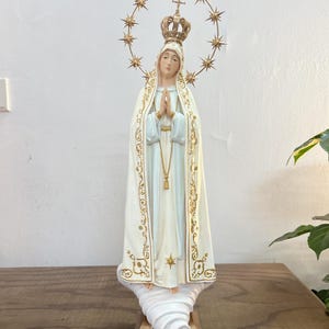 Könnte beinhalten: Religiöse Statue einer Frau in einem weißen Gewand mit goldenen Akzenten, einer Krone und einem Sternenhalo. Die Statue steht auf einer wolkenartigen Basis. Die Statue steht auf einer Holzoberfläche.