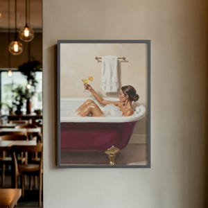 Könnte beinhalten: Ein gerahmter Druck zeigt eine Frau in einer Badewanne, die einen Cocktail hält. Die Badewanne mit Löwenfüßen ist tiefrot und mit Blasen gefüllt. Ein weißes Handtuch hängt an einem Holzhandtuchhalter. Das Kunstwerk hat einen Vintage-Stil.