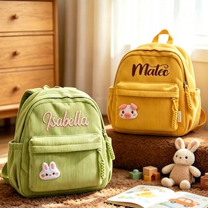 Puede incluir: Dos mochilas de pana, una verde con "Isabella" bordado y otra amarilla con "Mateo" bordado. Cada una tiene un bolsillo frontal y un parche de animalito. Un conejito de peluche y un libro infantil están en primer plano.