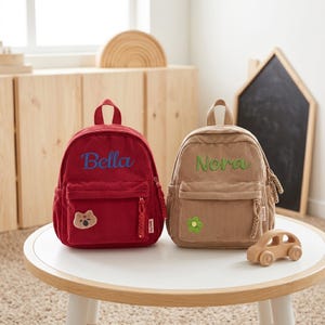 以下が含まれることがあります： 赤色の「Bella」と青色の刺繍、ベージュ色の「Nora」と緑色の刺繍が施された2つのコーデュロイバックパック。赤いバックパックにはクマのパッチが付いています。木製のおもちゃの車が近くにあります。バックパックは白いテーブルの上にあります。