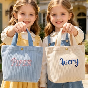 Könnte beinhalten: Zwei Cord-Tragetaschen, eine hellblaue mit rosa gesticktem "Piper", die andere beige mit blauem "Avery". Beide haben passende Griffe und einen Knopfverschluss. Die Taschen werden von zwei lächelnden Kindern gehalten.