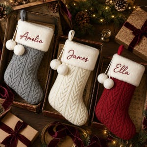 May include: Three Christmas stockings featuring a cable knit pattern in gray, cream, and red. Each stocking has a white faux fur cuff embroidered with the names Amelia, James, or Ella, and is decorated with two white pom-poms hanging from a cord.