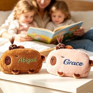 Op de afbeelding: Twee pluche etui's in de vorm van dieren, één bruin met "Abigail" geborduurd in groen, en één roze met "Grace" geborduurd in blauw. Elk heeft een stervormig detail en een kleine haarlok.