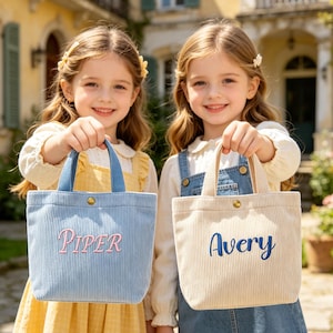 Könnte beinhalten: Zwei junge Mädchen halten Cord-Tragetaschen. Eine Tasche ist hellblau mit dem Namen "Piper" in Rosa gestickt. Die andere Tasche ist beige mit dem Namen "Avery" in Blau gestickt. Beide Taschen haben blaue Griffe und einen Knopfverschluss.