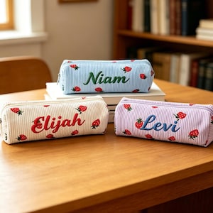 Op de afbeelding: Drie corduroy etuis in lichtblauw, crème en lavendel, elk geborduurd met een naam en aardbeienpatroon. De namen zijn "Niam", "Elijah" en "Levi". De etuis hebben een ritssluiting en liggen op een houten tafel.
