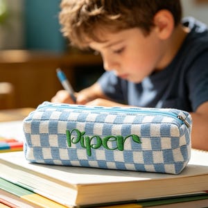 May include: A blue and white checkered pencil case with the name "Piper" embroidered in green script. The rectangular case has a blue zipper and is sitting on top of a stack of books. A child is in the background.