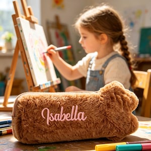 Könnte beinhalten: Ein braunes, flauschiges Federmäppchen mit dem Namen "Isabella" in Rosa gestickt. Das Federmäppchen liegt auf einem Holztisch mit bunten Markern. Im Hintergrund malt ein junges Mädchen an einer Staffelei.