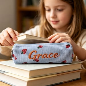 Könnte beinhalten: Hellblaue Stiftemäppchen aus Cord mit gestickten Erdbeeren und dem Namen "Grace" in orangefarbener Schrift. Das Federmäppchen hat einen Reißverschluss und liegt auf einem Stapel Bücher.