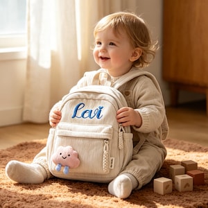 Könnte beinhalten: Cremefarbener Cord-Rucksack mit dem Namen "Lavi" in Blau gestickt. Der Rucksack hat eine kleine Vordertasche und eine wolkenförmige Applikation. Der Rucksack ist für Kleinkinder und junge Kinder konzipiert.