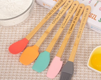 May include: Five-piece set of bamboo kitchen utensils with colorful silicone heads. Each spatula is engraved with a different phrase: "Taste of Heaven," "Mom Makes it Best," "Grandma's Kitchen," "Queen of the Kitchen," and "Life is What You Bake It."