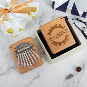 Può includere: Due kalimba in legno, una con lamelle metalliche e l'altra con il nome "ALEXANDER" inciso. Le kalimba sono su una superficie di marmo, una in una confezione regalo. Le kalimba sono di colore marrone chiaro.