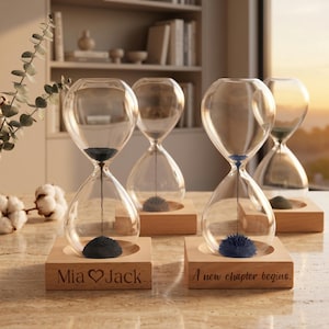 May include: Four clear glass hourglasses with magnetic sand on light wood square bases. Two bases feature engraved text: one reads "Mia ♡ Jack" and the other "A new chapter begins." The magnetic sand forms dark, spiky, sculptural shapes.