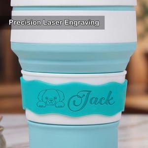 Close-up view focusing on the personalized laser engraving details of a lake blue collapsible silicone cup. On the white silicone band, a teal wave-shaped grip holds a cute engraved dog icon and the name "Jack" in a script font. This highlights the custom icon and name engraving option for pets.