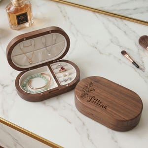 May include: A wooden jewelry box with an oval shape, open to reveal necklaces, rings, and a bracelet. The box is brown with a light-colored interior. The lid is closed and engraved with the name "Lillian" and floral accents. A perfume bottle and makeup brushes are in the background.