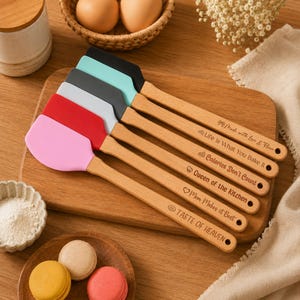 May include: A set of five silicone spatulas with wooden handles, each featuring a different color head including pink, red, white, gray, and teal. The handles are engraved with phrases such as Made with love & Flour, Life is What You Bake It, Calories Don't Count, Queen of the Kitchen, Mom Makes it Best, and TASTE OF HEAVEN.