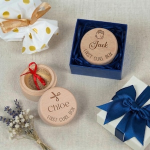 Puede incluir: Cajas de madera grabadas con los nombres "Jack" y "Chloe" y la inscripción "First Curl Box". Una de las cajas contiene un mechón de pelo. Otros regalos incluyen un regalo envuelto y una caja de regalo con una cinta azul.