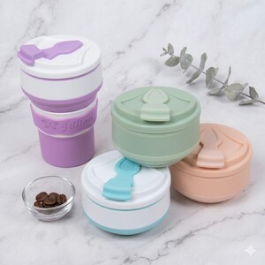 An aesthetic lifestyle set showing multiple personalized collapsible silicone cups on a kitchen marble surface. One extended purple cup features the name "Lillian" and a butterfly icon. Three other cups in light green, pale pink, and lake blue are folded into compact discs. A bowl of coffee beans and eucalyptus leaves complete the travel-ready scene.
