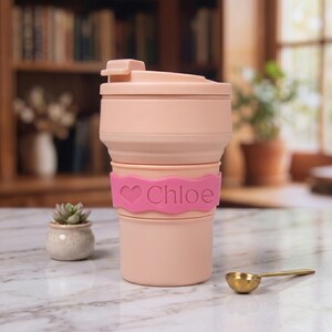 A close-up detailed view of the pale pink personalized collapsible silicone water cup. The vibrant neon pink name band displays an engraved heart icon and the name "Chloe." Set on a marble surface with a succulent and spoon, it emphasizes the feminine gift appeal.