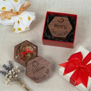 Puede incluir: Cajas de recuerdo hexagonales de madera, una abierta para revelar un mechón de cabello atado con una cinta roja. Una caja está en una caja roja y otra está abierta. Las cajas están grabadas con nombres y las palabras "First Curl Box."