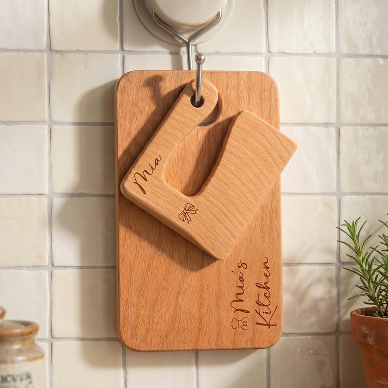 Personalized wooden kids knife and cutting board hanging neatly on a kitchen wall hook against white tiles. Engraved with Mia and a cute bow icon. This custom kids kitchen tool set doubles as beautiful decor and a Montessori fine motor skill toy.