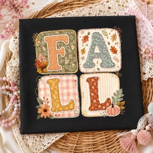 May include: A black t-shirt featuring a graphic design with the word FALL spelled out in four separate squares. Each letter is filled with a different autumn-themed pattern, including floral, plaid, and striped motifs, accented by leaves and pumpkins.